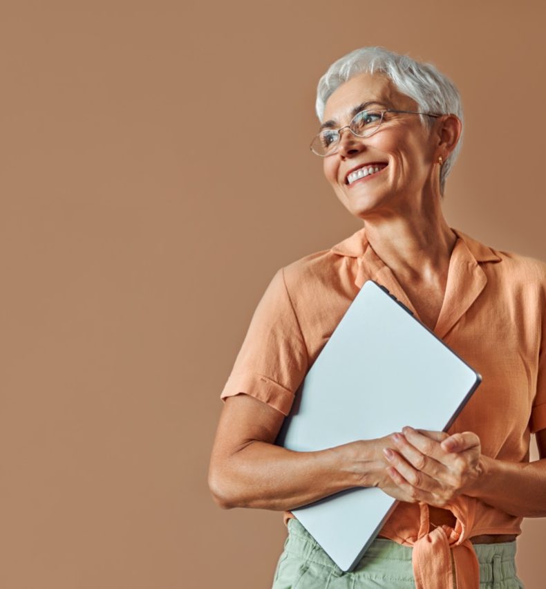 Confident beautiful mature gray haired stylish modern creative woman wearing glasses orange shirt and pastel green pants holding laptop and looking away to free space on beige background.